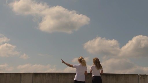 Zwei Frauen vor blauem Himmel, eine von ihnen zeigt nach links