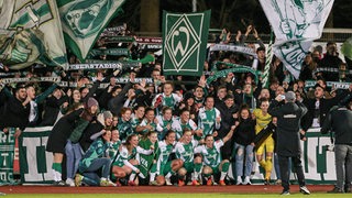 Werder-Spielerinnen kommen zum Gruppenfoto vor der Fan-Kurve der Ultras zusammen, die Fahnen und Schals schwenken.