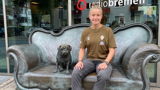 Werders Vizekapitänin Michelle Ulbrich sitzt lächelnd vor dem Radio-Bremen-Gebäude auf dem Loriot-Sofa neben dem Mops.