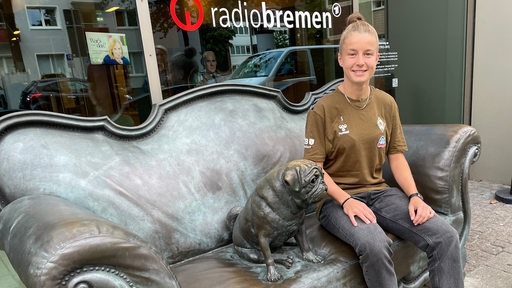 Werders Vizekapitänin Michelle Ulbrich sitzt lächelnd vor dem Radio-Bremen-Gebäude auf dem Loriot-Sofa neben dem Mops.