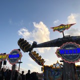 Das Fahrgeschäft "Airwolf" ist erleuchtet vor dem dunkler werdenen Himmel zu erkennen. Im Vordergrund sind Menschen als Schattenrisse zu erkennen.