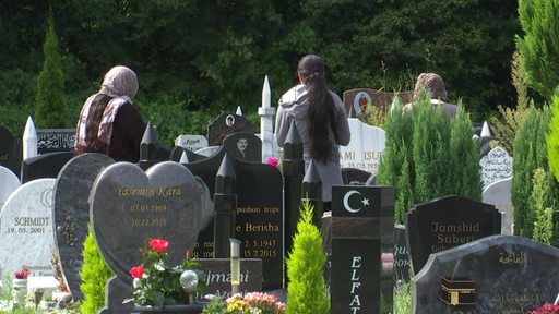 Drei Frauen stehen mit dem Rücken zur Kamera auf einem Friedhof vor einigen Gräbern. 