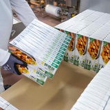 Eine Person in Arbeitskleidung hebt Verpackungen von einem Tisch.