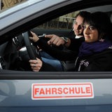 Eine junge Fahrschülerin sitzt neben dem Fahrlehrer, der ihr den Blick in den Seitenspiegel erklärt.