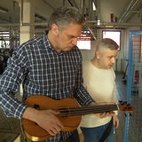 Zwei Männer stehen nebeneinander. Einer von ihnen spielt auf einer Ukulele.