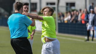 Ein Fußballspieler schubst auf dem Spielfeld den Schiedsrichter.