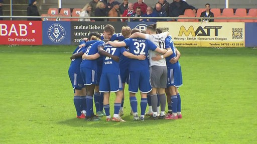 Auf dem Spielfeld stehen die Bremer SV Spieler in einem Kreis und umarmen sich.