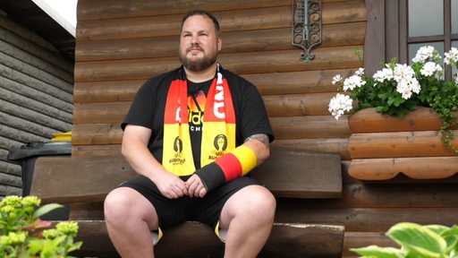 Ein Fußballfan sitzt mit Deutschland-Schal vor einer Holzhütte.