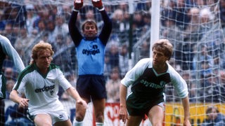 Die Fußball-Profis Winfried Schäfer von Gladbach und Klaus Fichtel von Werder Bremen sind im Pokal-Halbfinale 1984 im Zweikampf, hinter ihnen reckt sich Werder-Torwart Dieter Burdenski.