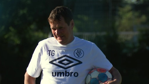 Der Trainer des Bremer SV Torsten Gütschow steht auf dem Fußballfeld mit einem Fußball unter dem Arm.