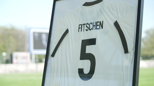 Ein weißes Fußballtrikot in einem Bilderrahmen, auf dem die Nummer 5 und Fitschen steht