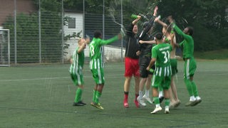 Fußballspieler von Tura Bremen hüpfen jubelnd und spritzen Sekt umher nach dem Gewinn der Meisterschaft.