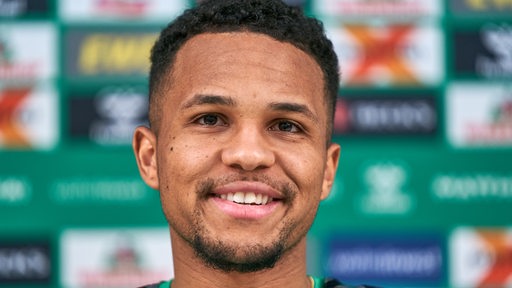 Werder-Profi Felix Agu lächelt auf dem Podium einer Pressekonferenz.