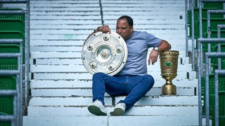 Werders Ex-Fußballer sitzt mit dem goldenen DFB-Pokal und der Meisterschale auf einer Treppe und küsst liebevoll die Schale.