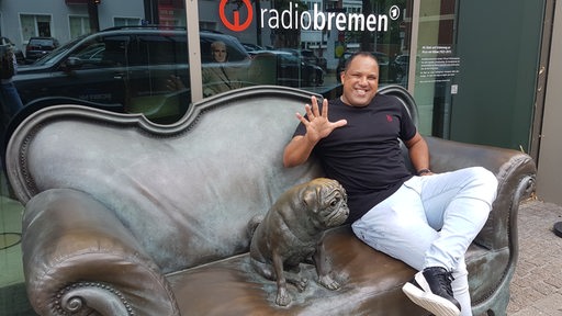 Ex-Fußball-Profi Ailton sitzt strahlend auf dem bronzenen Loriot-Sofa vor Radio Bremen und hält alle fünf Finger seiner rechten Hand hoch zu seinem 50. Geburtstag.