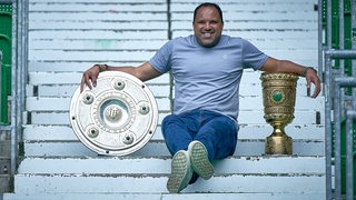 Ailton posiert sitzend auf einer Treppe in der Ostkurve des Weser-Stadion grinsend mit Meisterschale und DFB-Pokal.