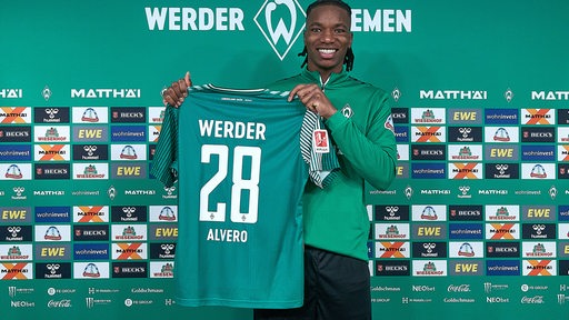 Werders Neuzugang Skelly Alvero hält strahlend bei seiner Vorstellung das grüne Trikot mit der Rückennummer 28 hoch.