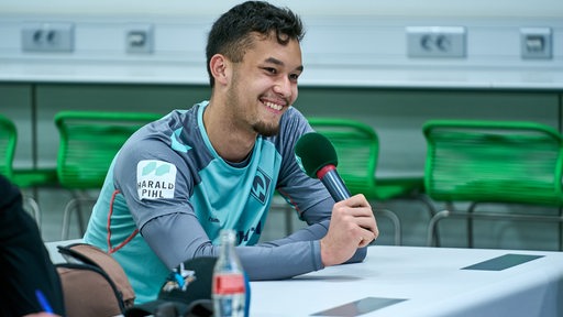 Werder-Torwart Mio Backhaus sitzt überraschend bei einer Pressekonferenz zwischen den Journalisten und stellt Keke Topp eine Frage.