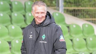Werders Geschäftsführer Frank Baumann steht lächelnd vor den grünen Schalensitzen der Tribüne im Stadion Platz 11.