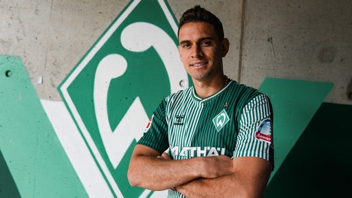 Werder-Neuzugang Rafael Borre posiert für ein Foto vor einer großen, aufgemalten Werder-Raute an der Wand.