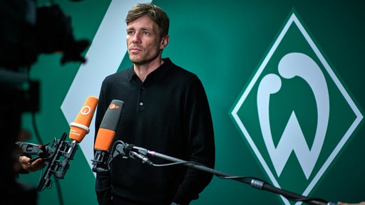 Werder-Geschäftsführer Clemens Fritz steht vor einer grünen Wand mit weißer Werder-Raute vor einer Kamera beim Interview.