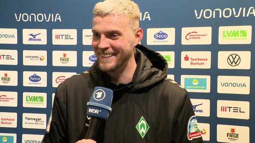 Werder-Stürmer Marvin Ducksch steht lächelnd nach dem Spiel in Bochum vor einer Werbewand beim Interview.
