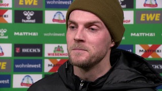 Werder-Stürmer Marvin Ducksch trägt eine dunkelgrüne Wollmütze und steht nach einem Spiel vor einer Werbewand beim Interview.