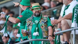 Kultiger Werder-Fan mit Hut, Kutte und Sonnenbrille steht mit Bierbecher erwartungsfroh im Fan-Block.