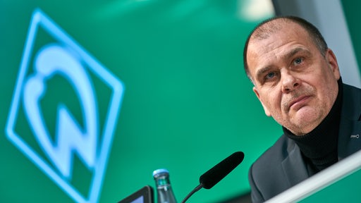 Werder-Finanzchef Klaus Filbry nachdenklich auf dem Podium einer Pressekonferenz, im Hintergrunde eine weiße Werder-Raute an der grünen Wand.