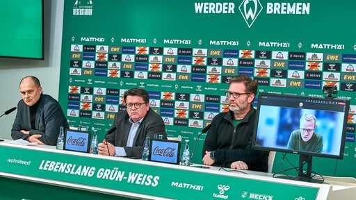 Werder-Finanzchef Filbry, Präsident Hess-Grunewald, Pressesprecher Pieper sitzen auf dem Podium einer Pressekonferenz, Aufsichtsratsmitglied Ohlmeyer ist auf einem Bildschirm zugeschaltet.