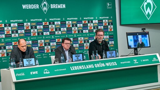 Werder-Finanzchef Filbry, Präsident Hess-Grunewald, Pressesprecher Pieper sitzen auf dem Podium einer Pressekonferenz, Aufsichtsratsmitglied Ohlmeyer ist auf einem Bildschirm zugeschaltet.