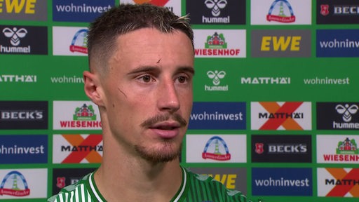 Werder-Kapitän Marco Friedl steht nach dem Sieg gegen Mainz vor einer Werbewand beim Interview.