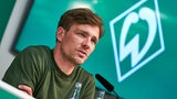 Werders Sportlicher Leiter Clemens Fritz sitzt auf dem Podium einer Pressekonferenz, im Hintergrund ein Bildschirm mit einer weißen Werder-Raute auf grünem Untergrund.