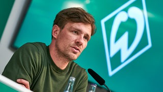 Werders Sportlicher Leiter Clemens Fritz sitzt auf dem Podium einer Pressekonferenz, im Hintergrund ein Bildschirm mit einer weißen Werder-Raute auf grünem Untergrund.
