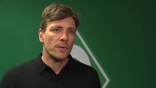 Werder-Geschäftsführer Clemens Fritz steht vor einer grünen Wand mit der weißen Werder-Raute bei einem Interview.