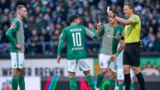 Ein Schiedsrichter zeigt Werder-Spieler Leonardo Bittencourt die Gelbe Karte, Marvin Ducksch und Rafael Borré stehen daneben.