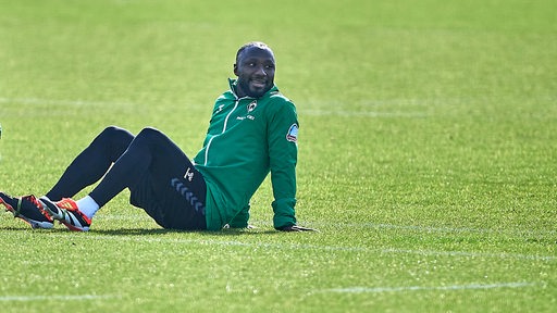 Werder-Spieler Naby Keita liegt am Rande des Trainings auf dem Rasen im Sonnenlicht und beobachtet seine Mitspieler.