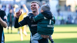 Werder-Stürmer Marvin Ducksch hält nach dem Sieg in Kiel seinen Sohn auf dem Arm und winkt den Bremer Fans zu.