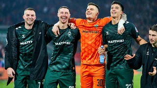 Die Werder-Spieler Ducksch, Friedl, Zetterer und Malatini stehen fröhlich Arm in Arm nebeneinander und feiern mit den Fans den Sieg gegen Köln.