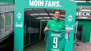 Werder-Neuzugang Dawid Kownacki hält vor dem Spielertunnel lächelnd sein Werder-Trikot mit der Nummer neun hoch.