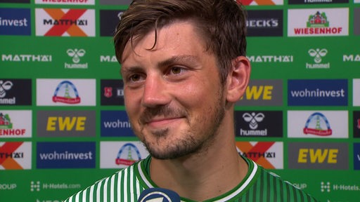 Werder-Stürmer Dawid Kownacki lächelt vor einer Werbewand nach dem Sieg gegen Mainz bei einem Interview.