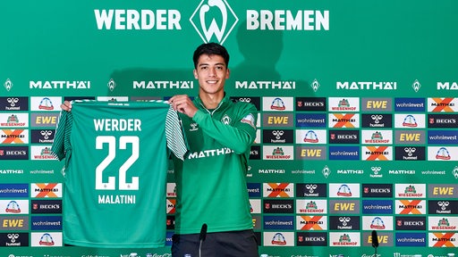 Werder-Neuzugang Julian Malatini hält vor der Pressekonferenz strahlend sein Werder-Trikot in die Kameras.
