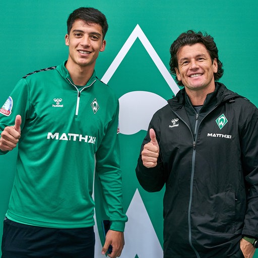 Werder-Neuzugang Julian Malatini posiert lächelnd mit Co-Trainer Nelson Valdez vor einer großen Werder-Raute an der Wand.