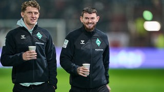Die Werder-Fußballprofis Jens Stage und Oliver Burke schlendern entspannt und lächelnd vor einem Spiel mit einem Kaffeebecher in ihren Händen über das Spoielfeld.