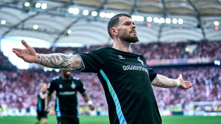 Werder-Stürmer Oliver Burke steht nach seinem Tor gegen Stuttgart stolz und mit weit ausgebreiteten Armen vor dem Gästeblock der Werder-Fans.