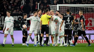 Werder-Spieler um Kapitän Marco Friedl umringen aufgebracht Schiedsrichter Dankert beim Spiel in Stuttgart.