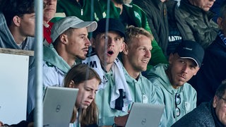Bei einem Testspiel in Meppen sitzen Werder-Kapitän Marco Friedl und seine Mitspieler Jens Stage, Marco Grüll und Senne Lynen auf der Tribüne zwischen Zuschauern und Journalisten an Laptops.