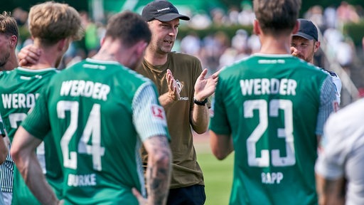 Werder-Trainer Ole Werner ist am Spielfeldrand umringt von seinen Spieler und erklärt ihnen etwas.