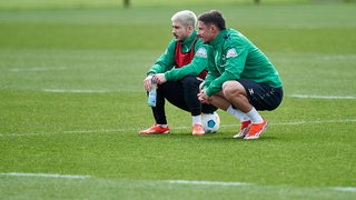 Die Werder-Spieler Marco Friedl und Romano Schmid hocken am Rande des Trainings auf dem Rasen und schauen in die gleiche Richtung.