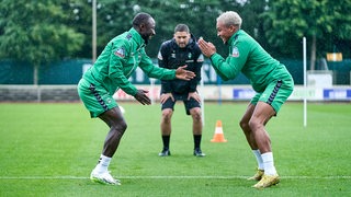 Die Werder-Profis Naby Keita und Felix Agu stehen sich beim launigen Einzeltraining gegenüber bei einer Übung und klatschen ab.